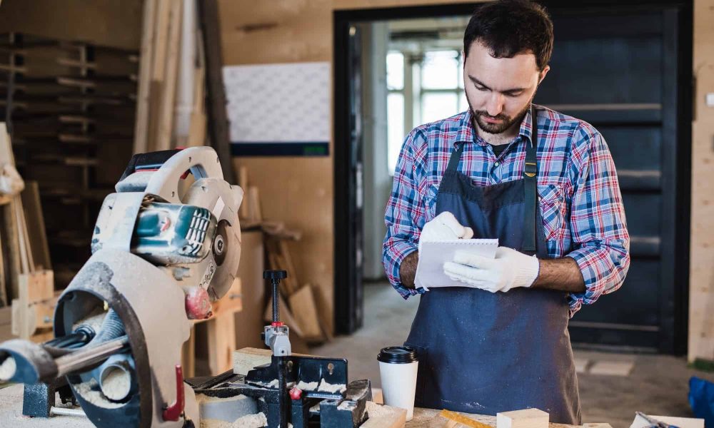 Carpenter taking a coffee break holding notebook in front of circular saw at his workshop